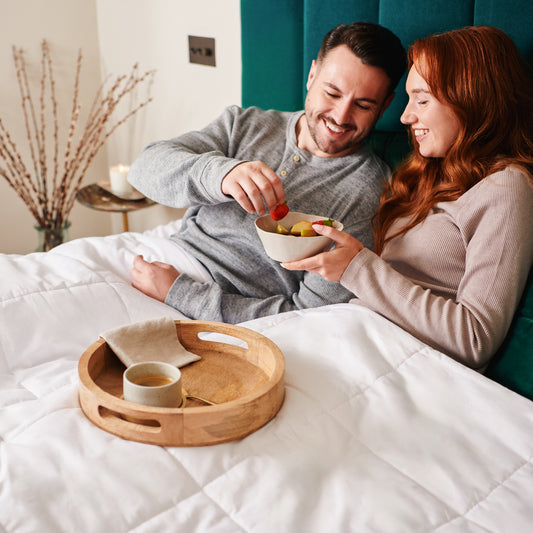 A couple sit up in bed covered by their heated duvet eating fruit in bed